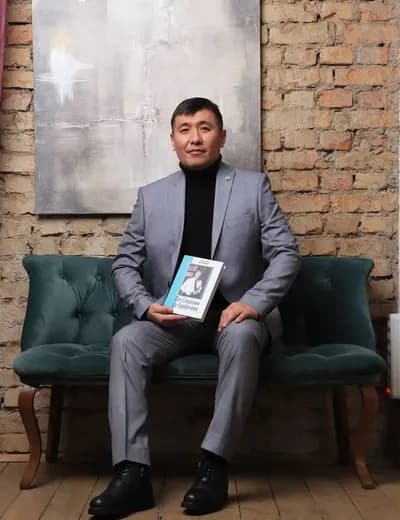 Қалдарбек Сраилов кірпіш қабырғаның алдындағы орындықта отыр, қолында кітап